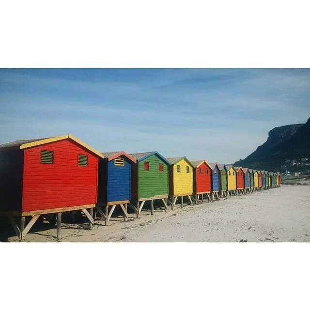A row of colorful beach huts on stilts along sandy Muizenberg beach with a mountain in the background.