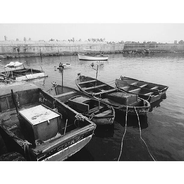 Black and white photo of small boats docked at a harbor with a stone wall and ships in the background.