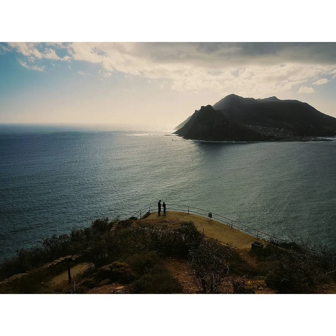 Two people standing on a grassy cliff overlooking the ocean with a mountain in the background