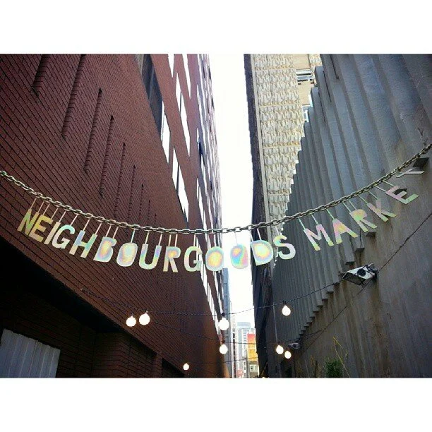 String of colorful letters spelling "NEIGHBOURGOODS MARKET" hanging between tall buildings with string lights