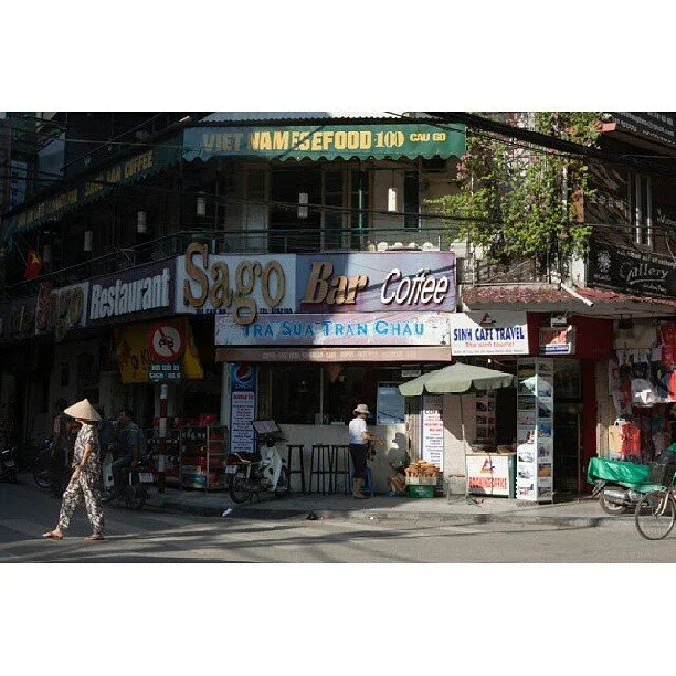 Street view with a Vietnamese restaurant and coffee shop, signs for Saigon Bar and Coffee, and a street vendor with an umbrella.