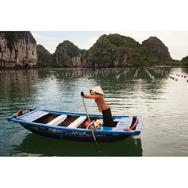 A person standing on a small blue boat in a body of water, using a pole to steer. The person wears a conical hat and brown jacket. In the background are large, green, rocky islands and buoys floating in the water.