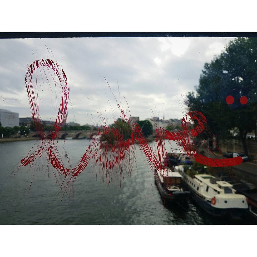 Window with red marker writing and scratches, overlooking a river with boats and buildings, cloudy sky, trees on the right.