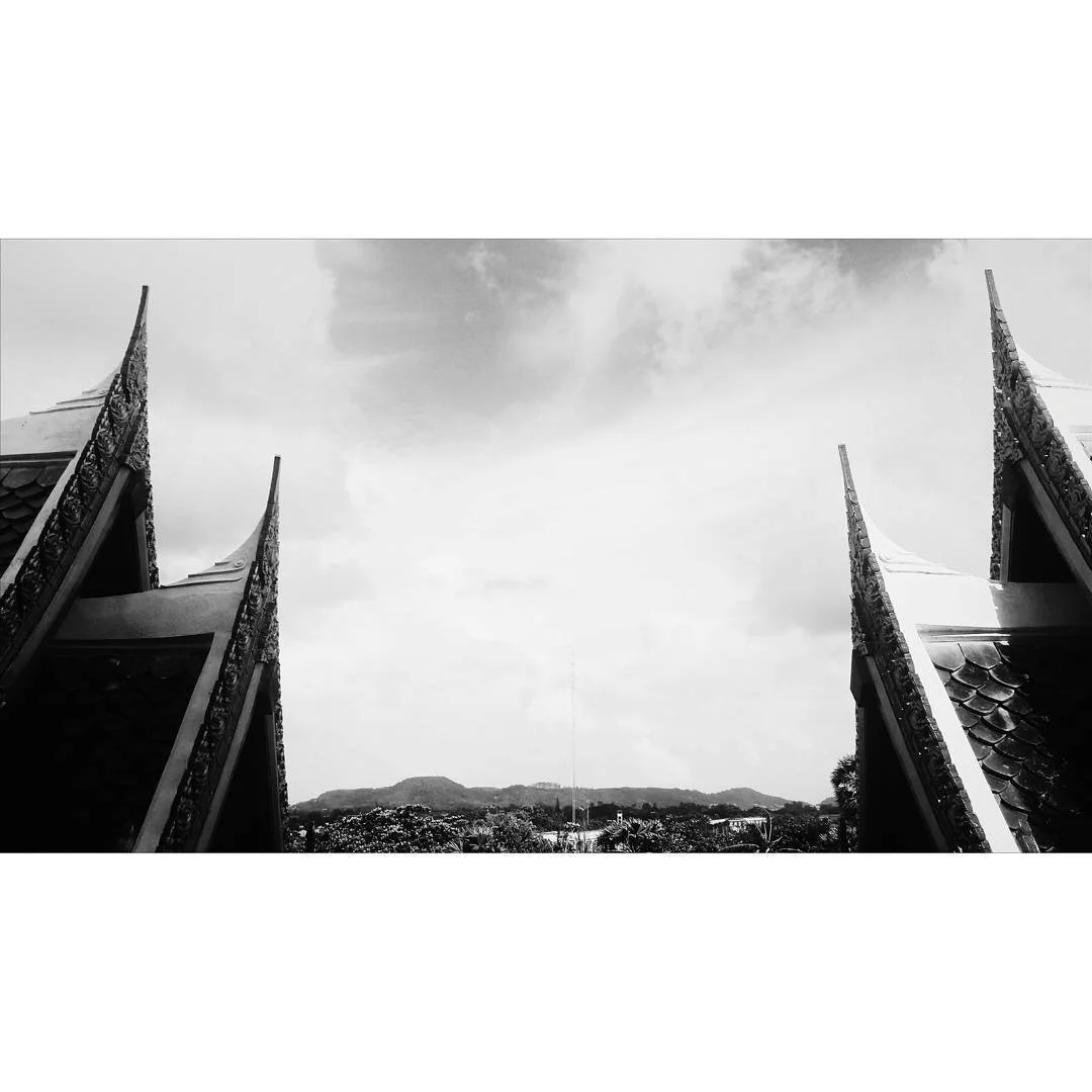 Black and white photo of traditional Thai temple rooftops with ornate decorations, framing a view of distant hills and cloudy sky.