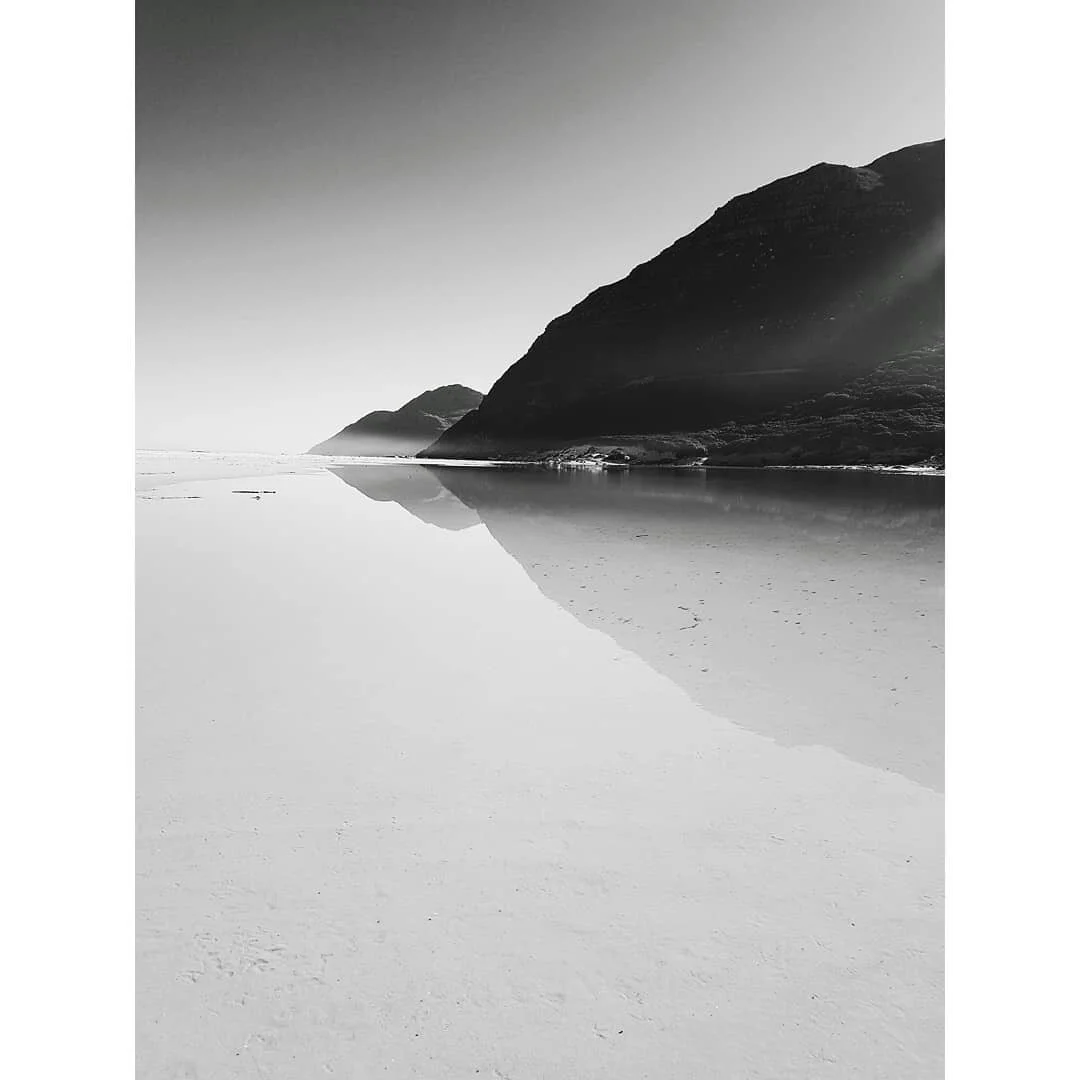Black and white photo of a beach with calm water reflecting mountains in the background.