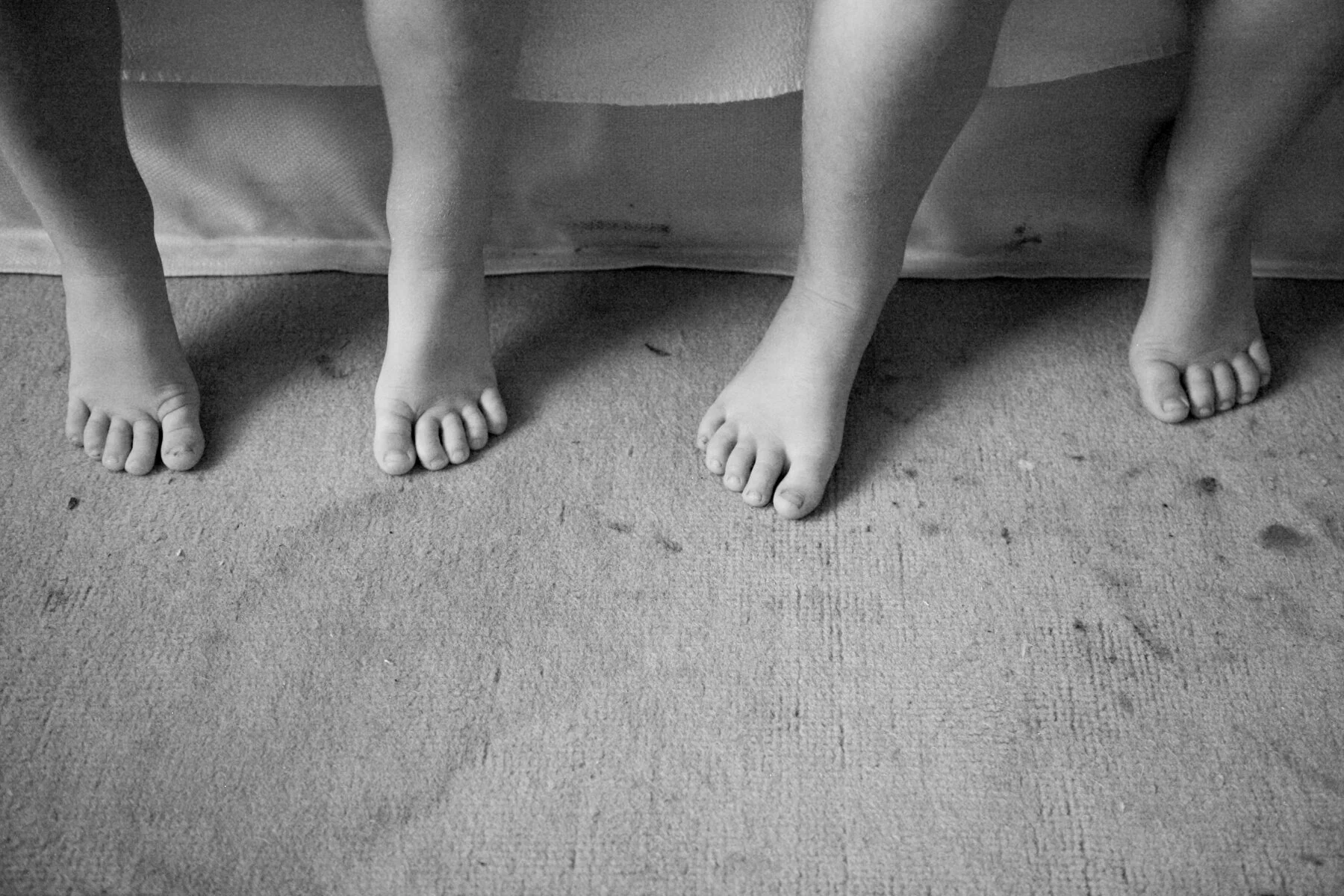 Close-up of three children's feet standing on a textured carpeted floor, with a cloth or blanket hanging down in the background.