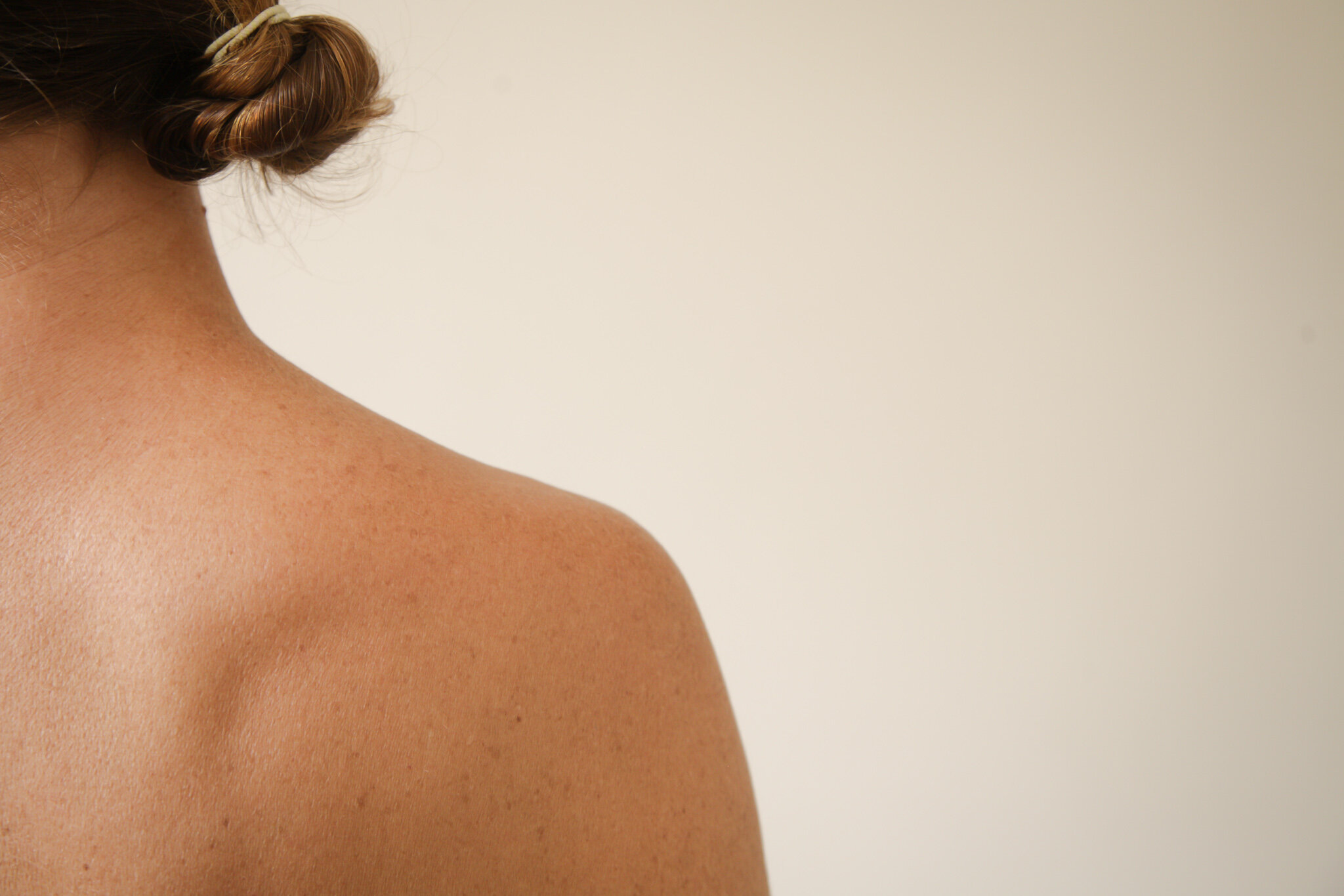Close-up of a person's shoulder and neck with light skin tone, brown hair tied up, against a plain background.