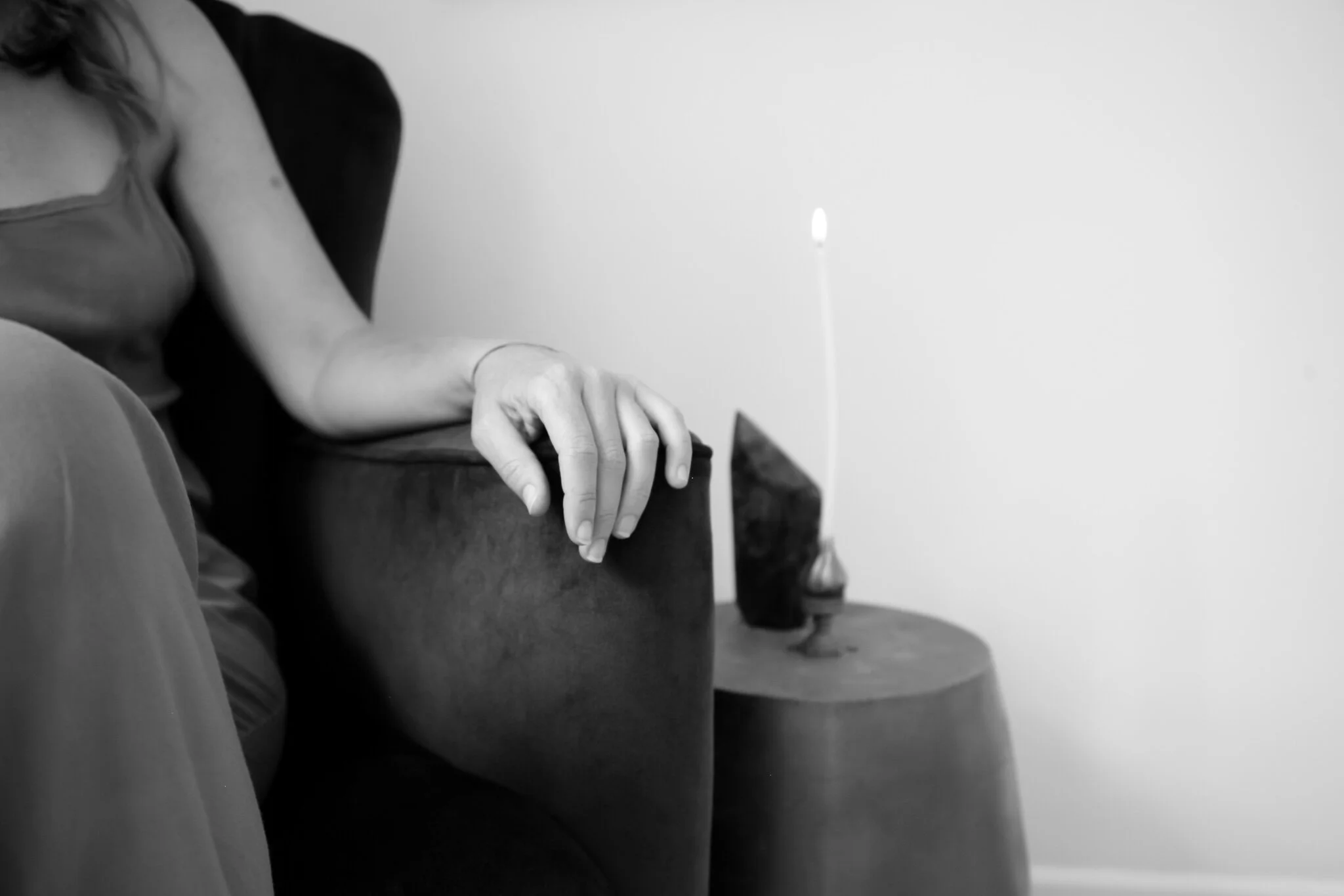 Close-up of a person's hand resting on an armrest of a dark sofa in a black-and-white photograph. There is a small wooden table beside the sofa with a decorative object and a lit candle.