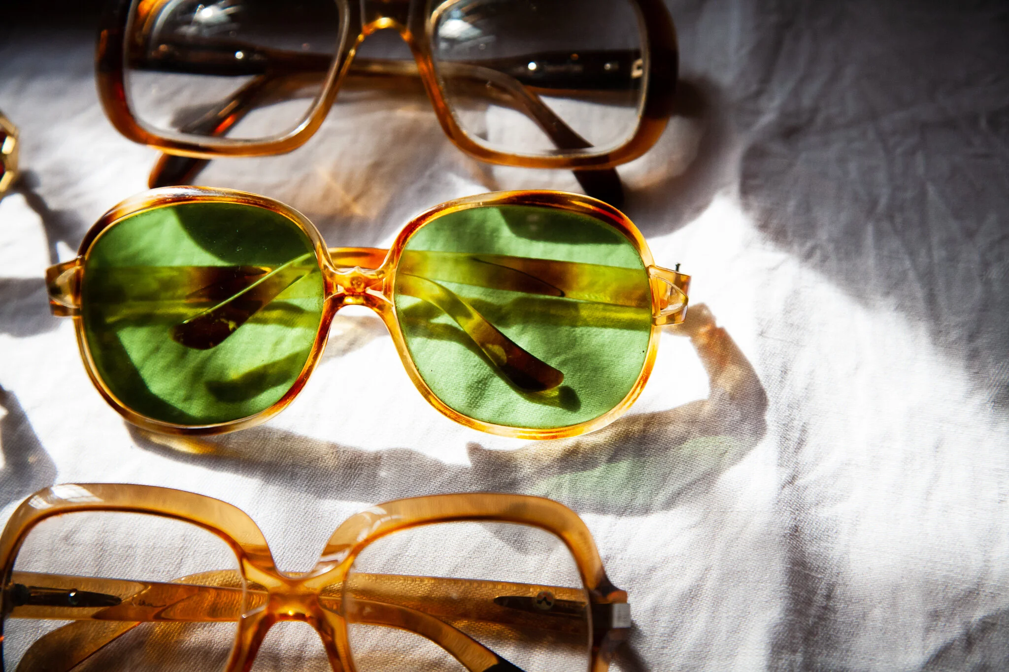 Collection of sunglasses with different styles and colors on a white fabric surface, with sunlight casting shadows.