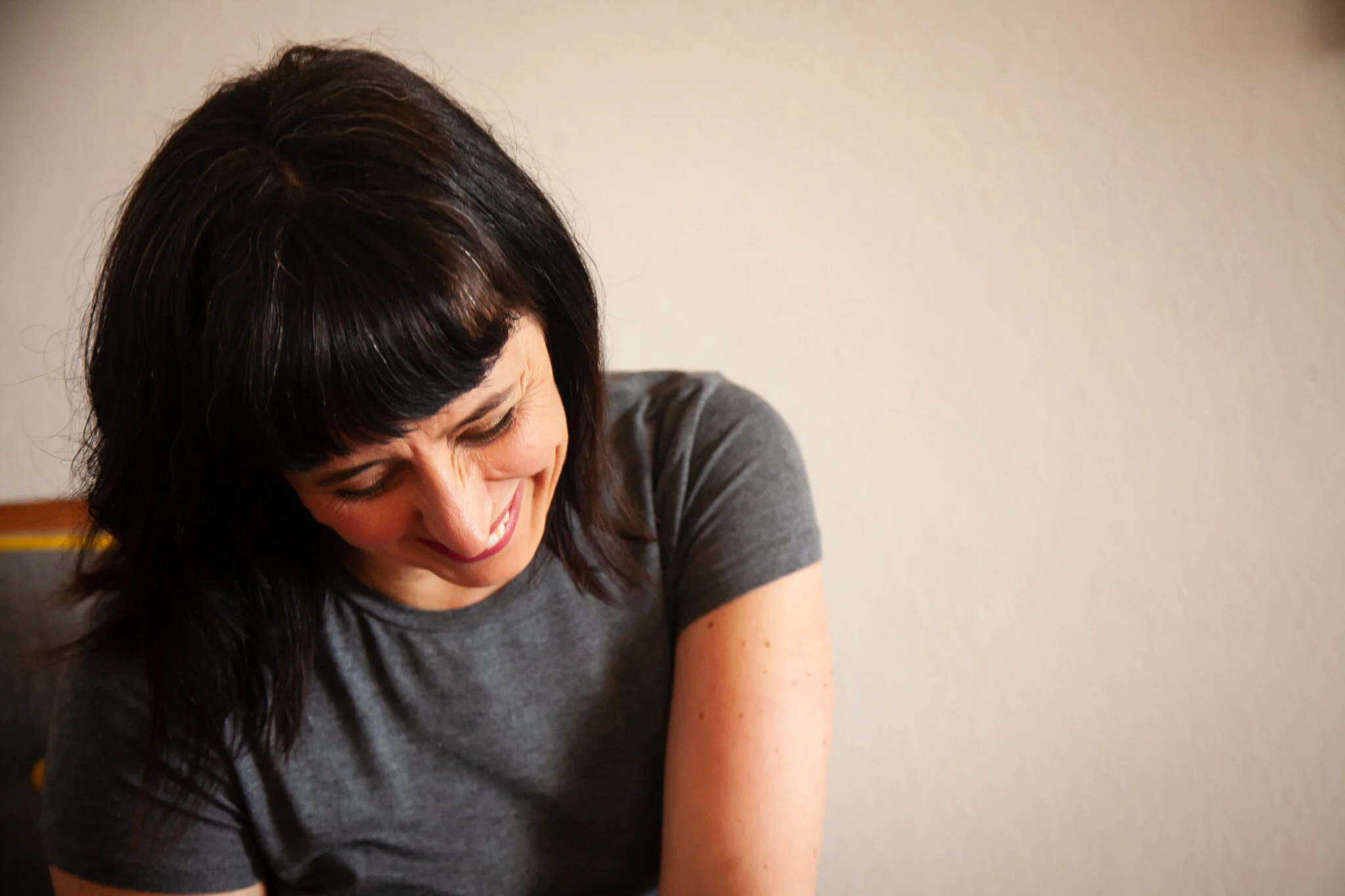 A woman with dark hair and bangs smiling and looking down, wearing a gray t-shirt, with a plain light-colored wall in the background.
