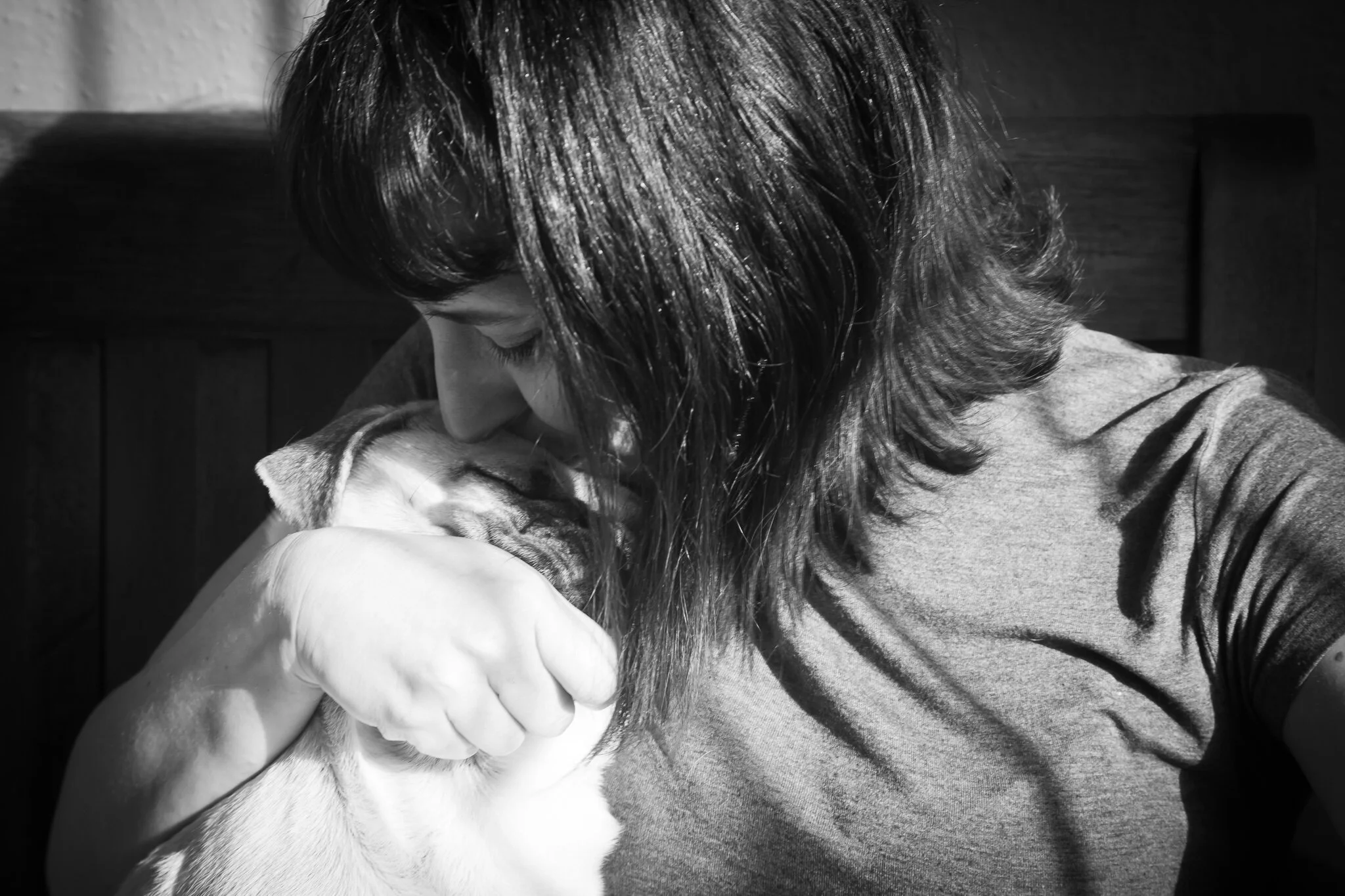A woman with shoulder-length hair in a grey shirt is kissing a dog with closed eyes while holding it close. The dog is a Pug and is resting peacefully.