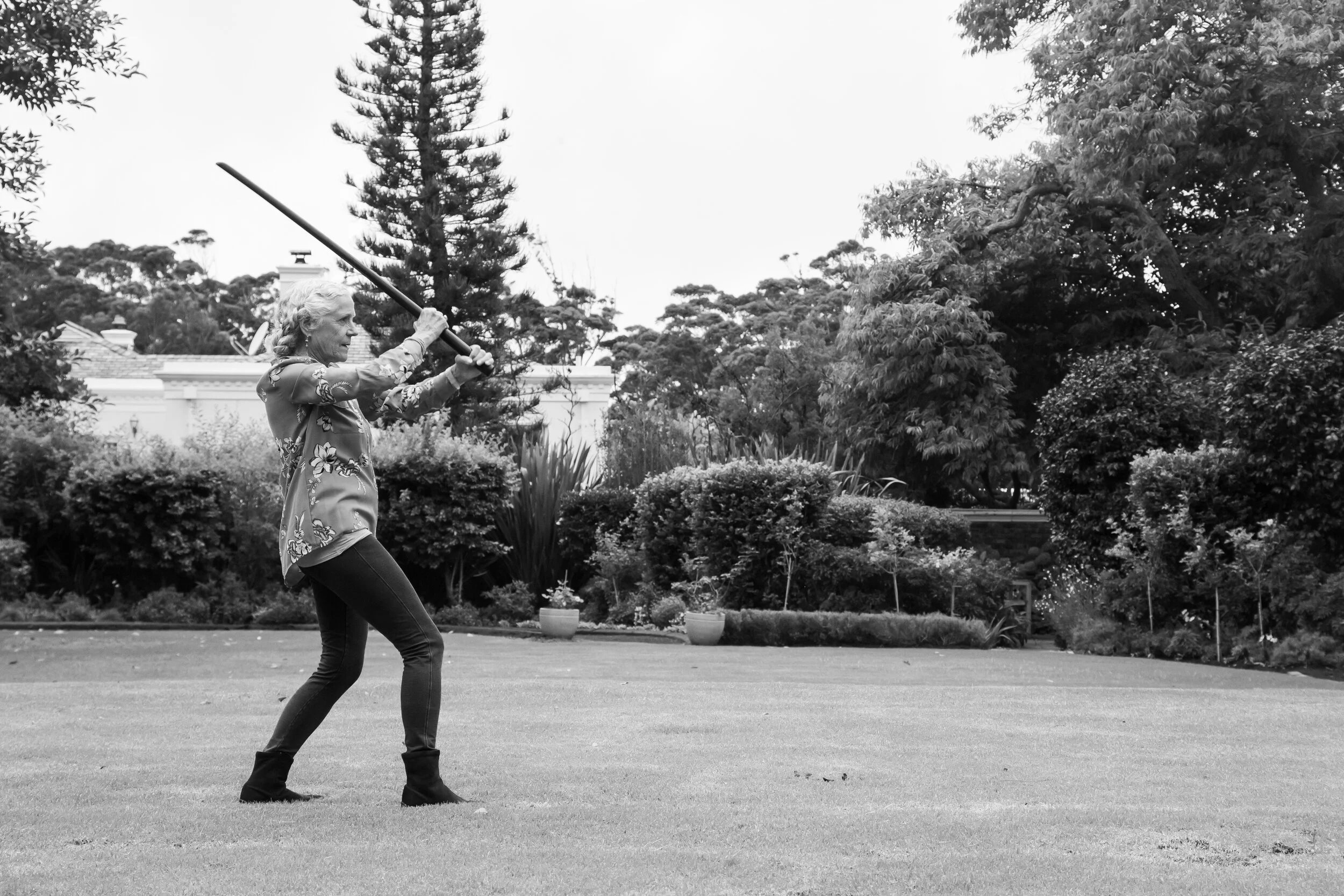 A woman in a floral button-up shirt and dark pants is standing on a well-maintained lawn, holding a sword with both hands in a practice stance. She appears to be outdoors in a garden with trees, bushes, and potted plants in the background.
