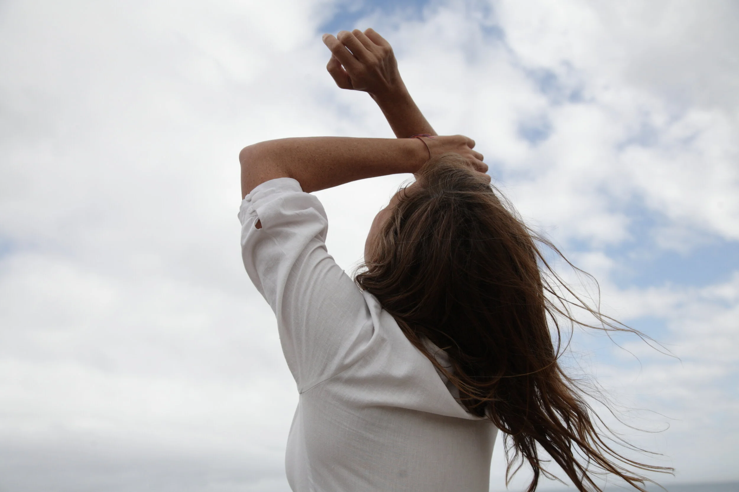 A woman with brown hair wearing a white shirt outdoors against a cloudy sky, with her arms raised and her hand touching her head.
