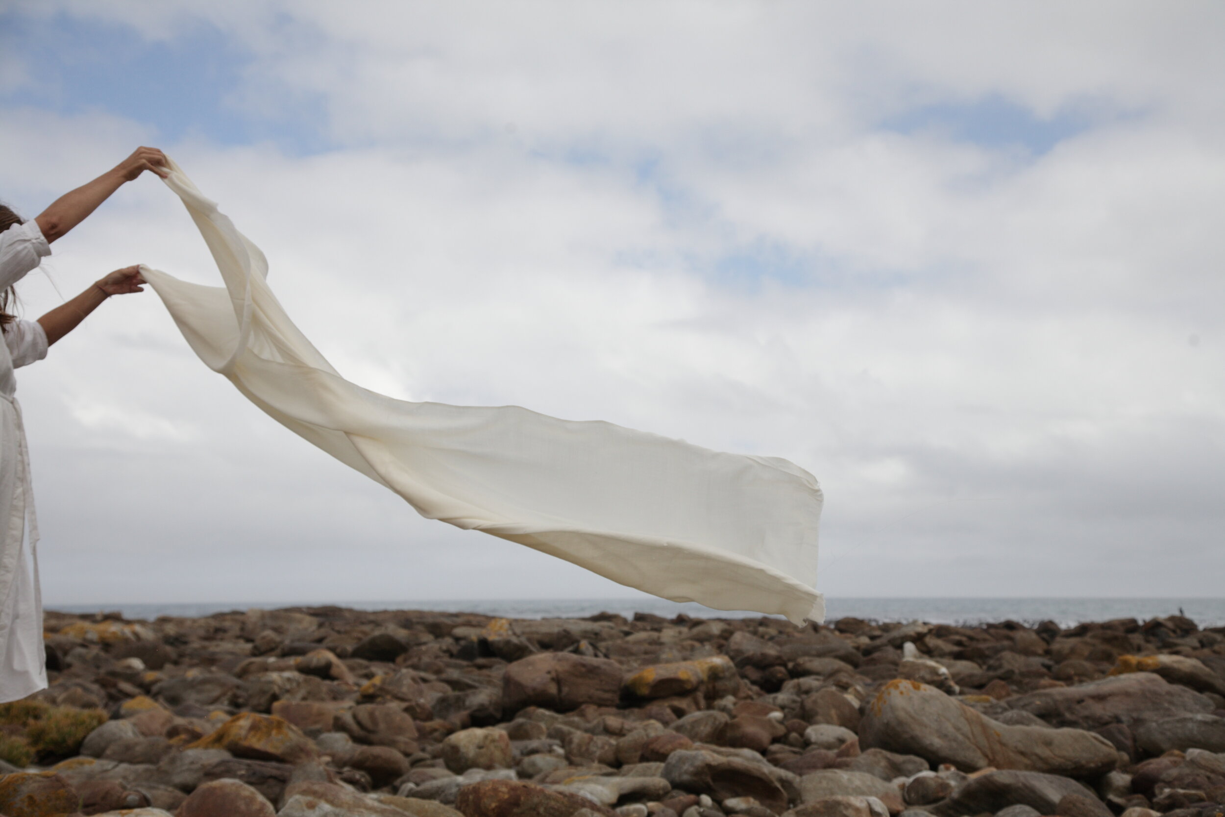 Two women with long hair holding and waving a white fabric or sheet on a rocky beach with cloudy sky in the background.