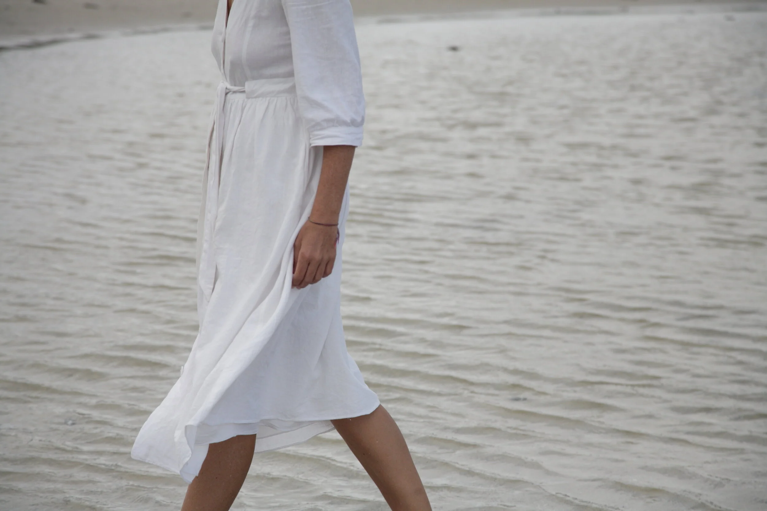 Person walking on the beach near the water, wearing a white dress.