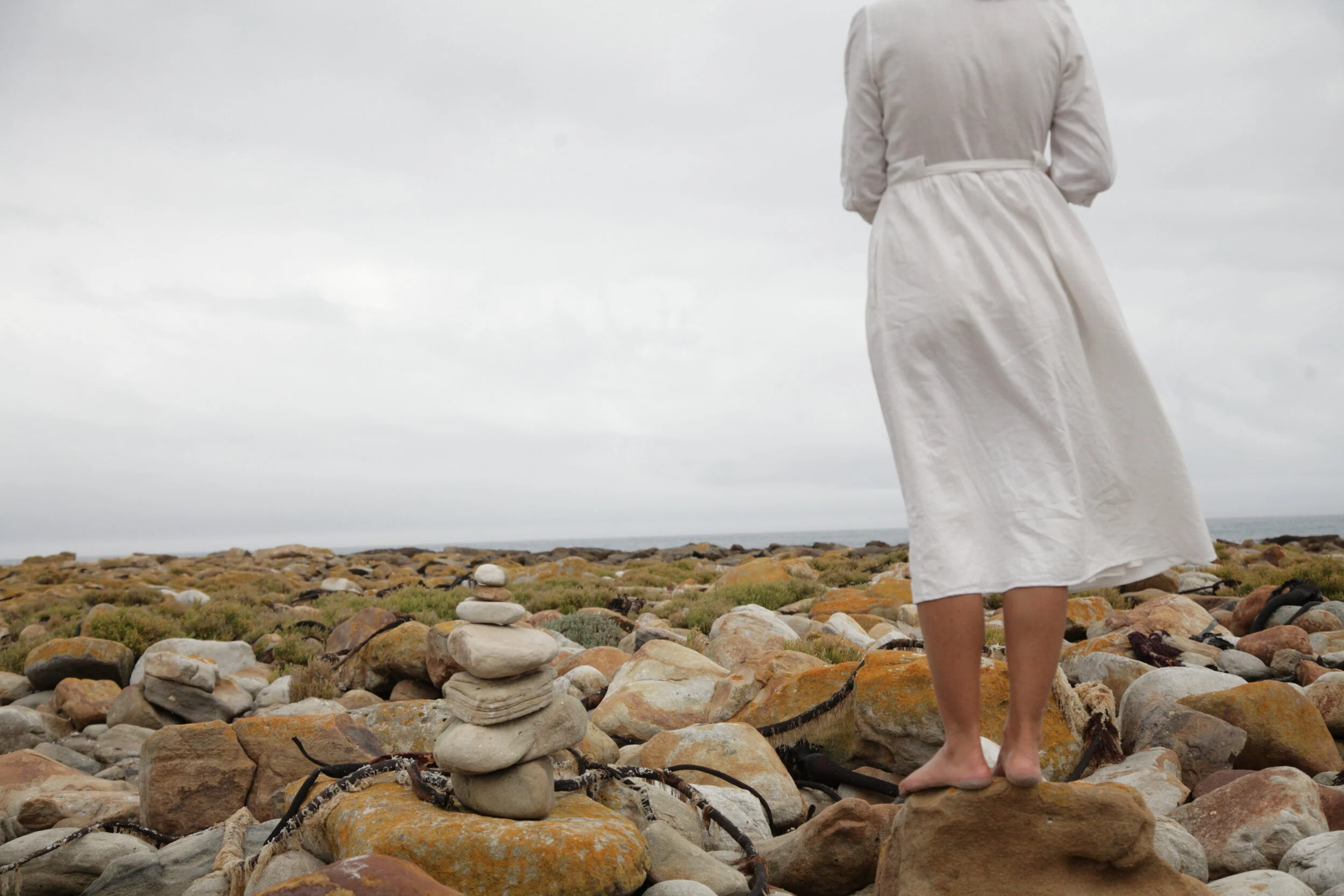Person in a white dress standing on rocks near the coast, with stacked rocks in the foreground and cloudy sky above.