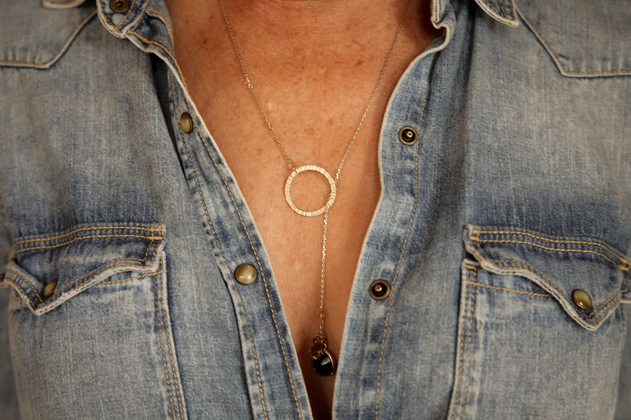 Close-up of a person's chest wearing a denim shirt unbuttoned, showing layered gold necklaces with circular and teardrop pendants.