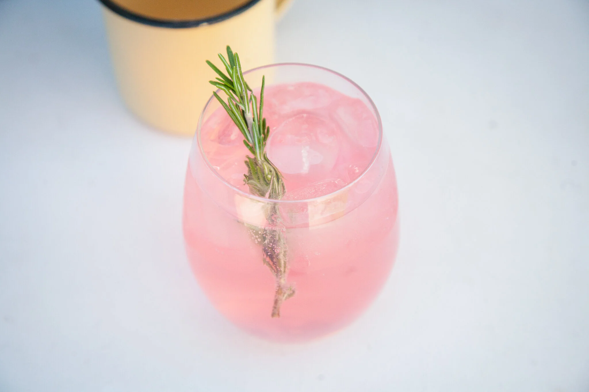 A pink drink with ice and a sprig of rosemary in a clear glass, with a yellow container in the background on a white surface.