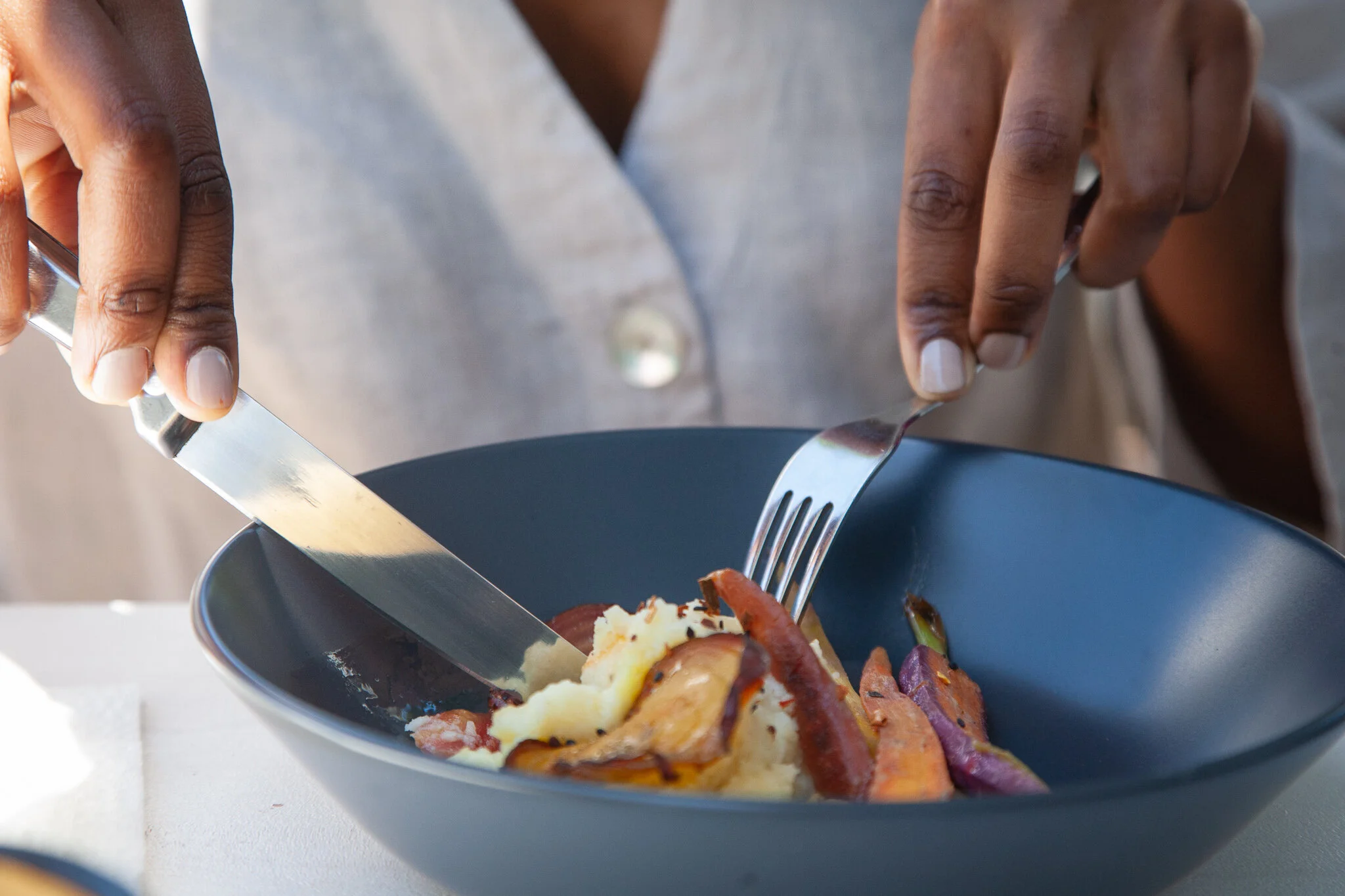 A person is using a knife and fork to eat a dish that includes mashed potatoes, roasted carrots, and bacon in a dark bowl.