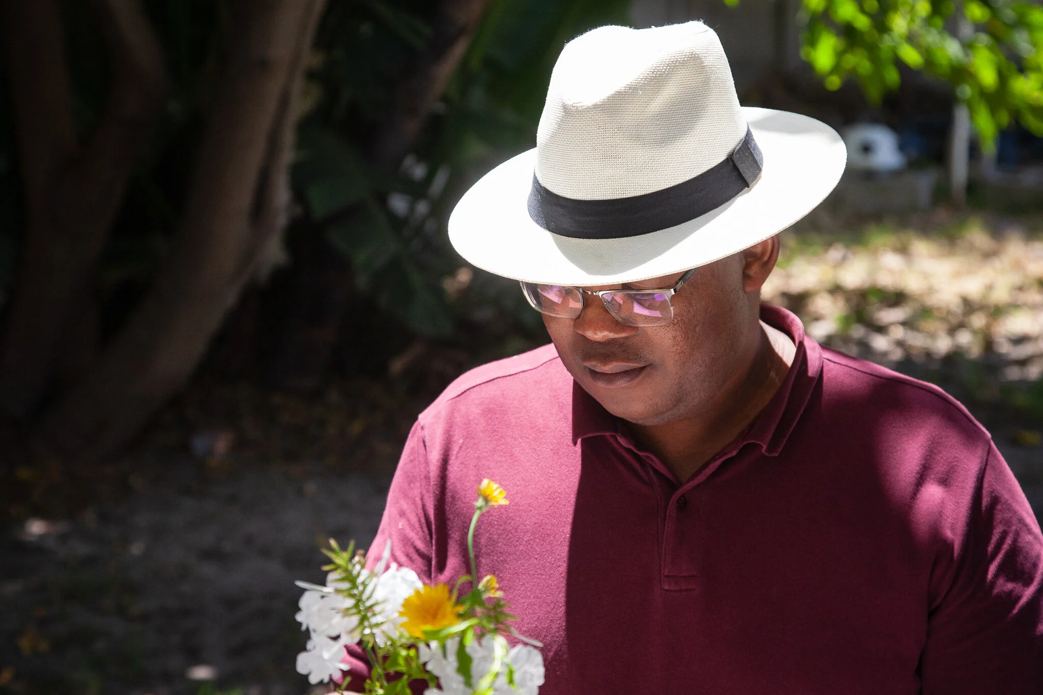 A man wearing a white fedora with a black band, glasses, and a maroon shirt, looking down at a bouquet of wildflowers outdoors in sunlight.