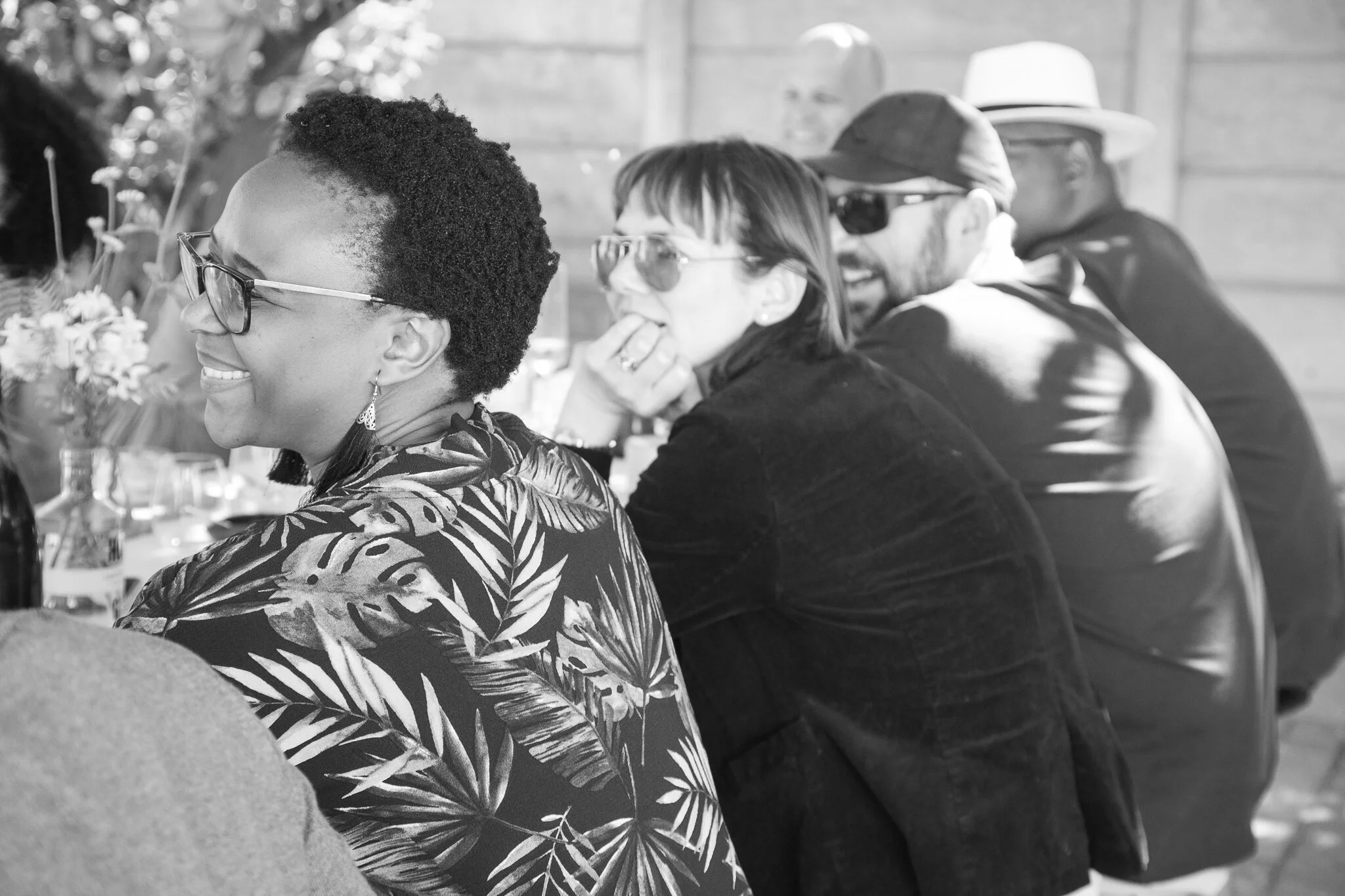 Group of four people sitting at a table, smiling and talking at an outdoor gathering, with flowers and bottles on the table.