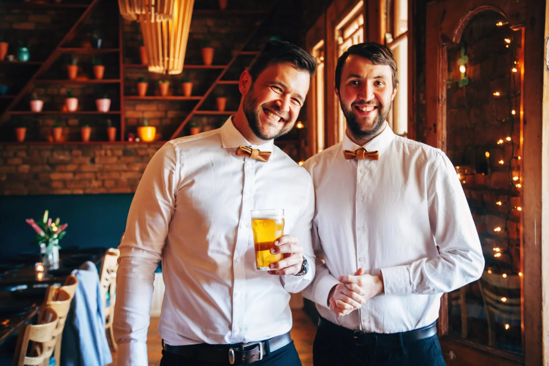 Two smiling men in white shirts and bow ties, one holding a drink, standing inside a warmly lit restaurant with brick walls and decorative shelving.