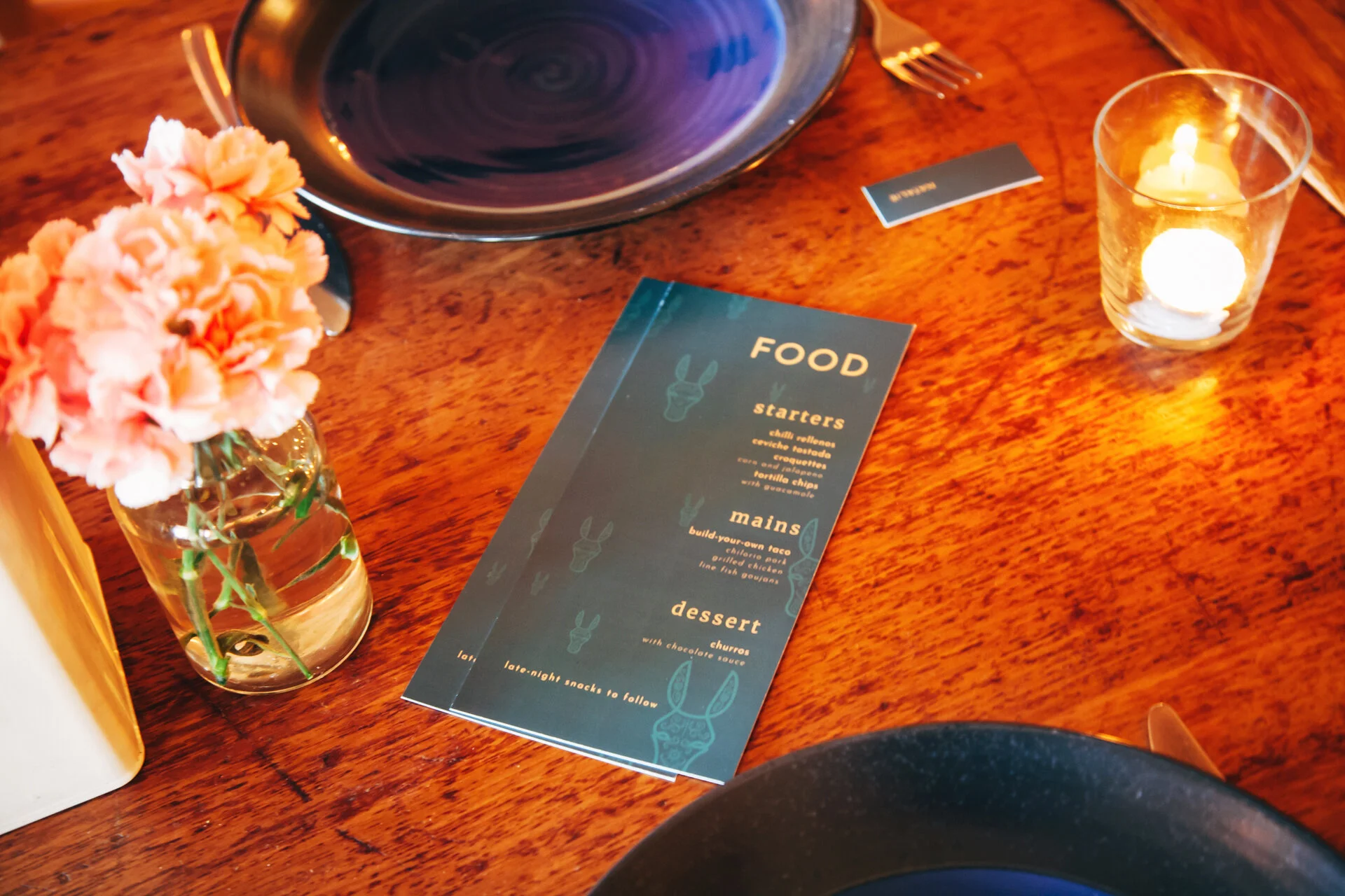 A wooden table set for a meal with a black plate, a menu card, a small card, a glass candle holder with a lit candle, and a vase of pink flowers.