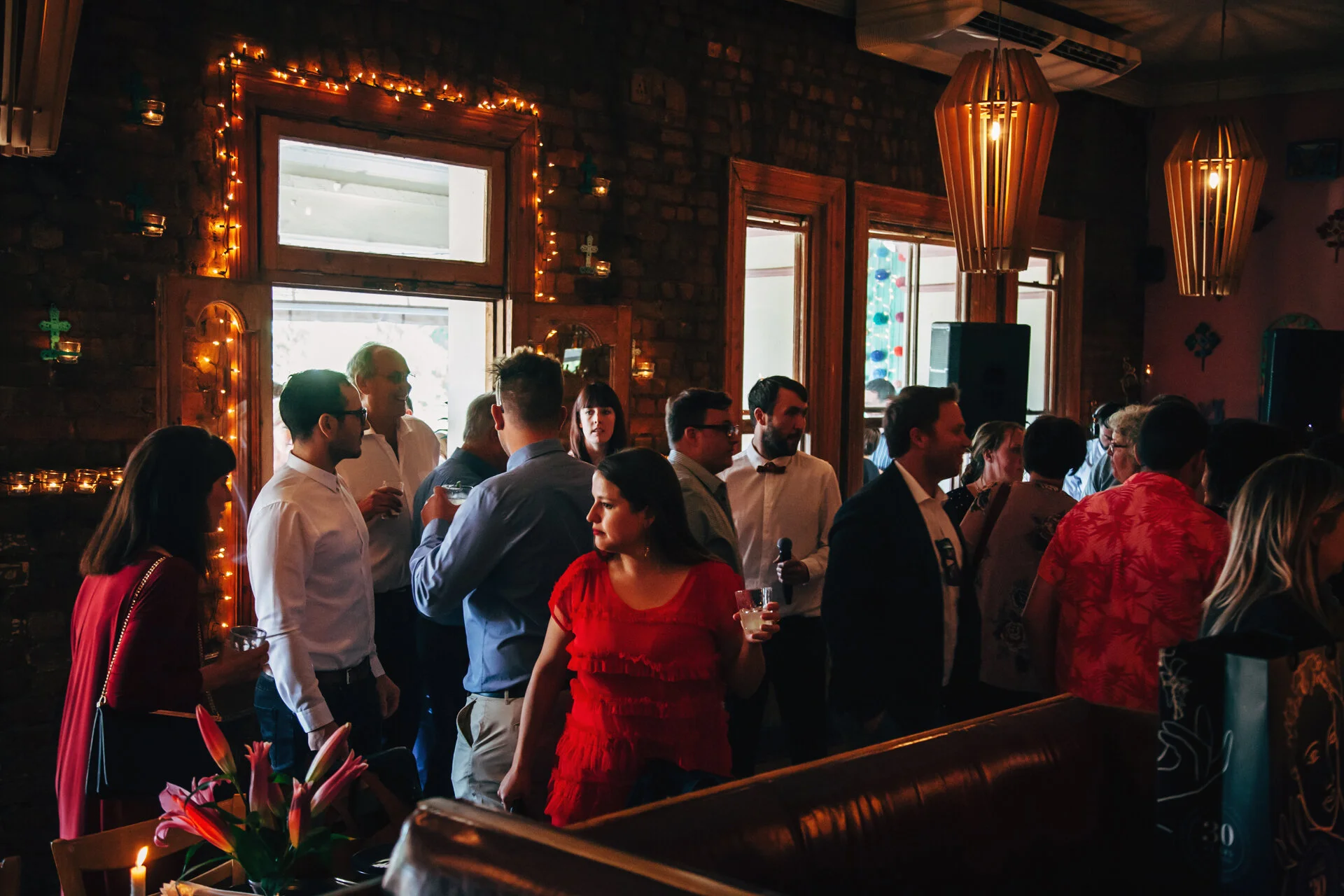People gathered in a dimly lit indoor party or event space, engaging in conversation, with warm lighting, wooden accents, and decorative lighting fixtures hanging from the ceiling.