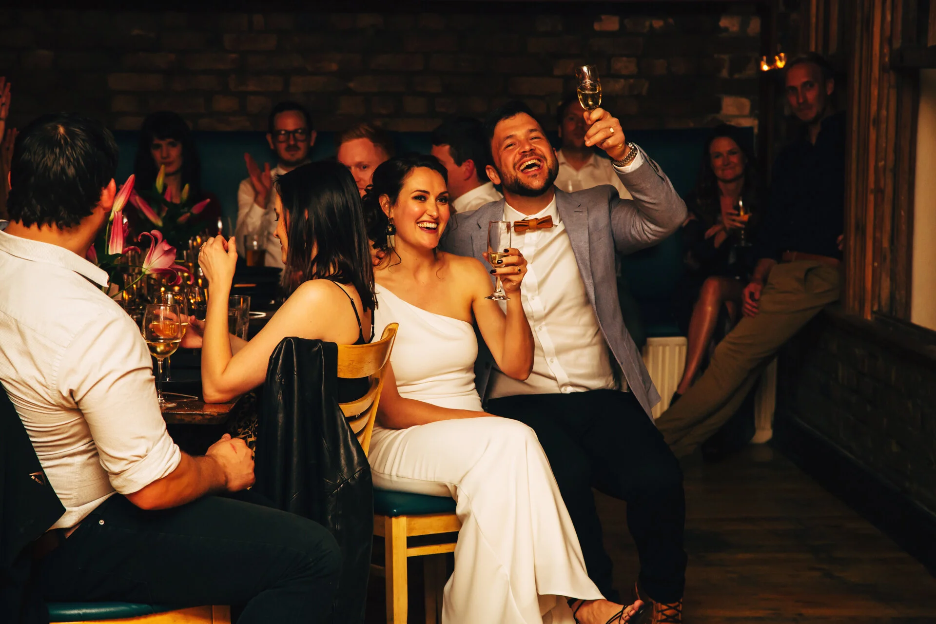 People at a celebration or party, smiling and raising glasses of champagne in a warmly lit room with brick walls.