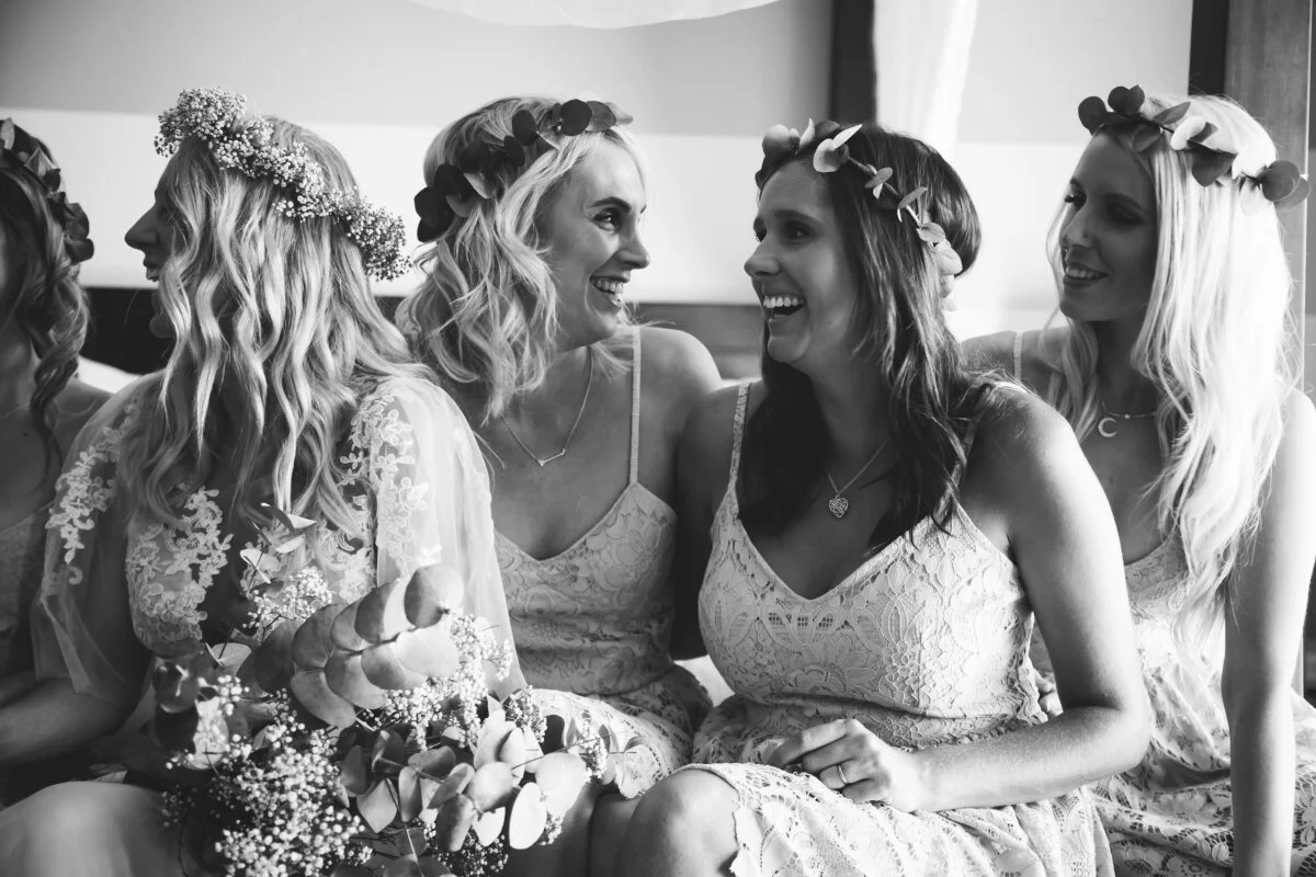 Black and white photo of five women sitting together, wearing flower crowns, smiling, and dressed in lace dresses, celebrating a wedding or bridal event.