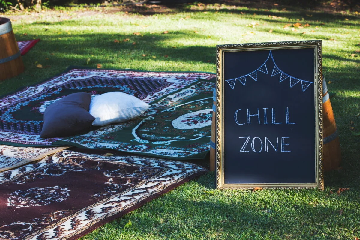 An outdoor chill zone setup with rugs and pillows on grass, featuring a chalkboard sign that reads 'Chill Zone' decorated with a triangle banner.
