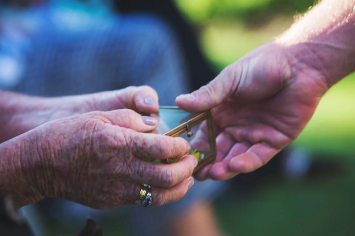 Close-up of two hands, one elderly and one younger, exchanging a small glass box with a gold frame outdoors.