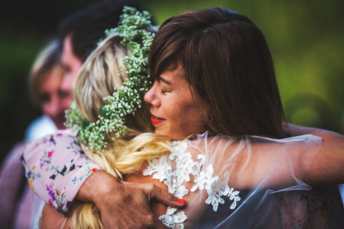 Women hugging at a wedding, one wearing a floral dress and a floral headband, the other in a wedding dress, with a blurred background.
