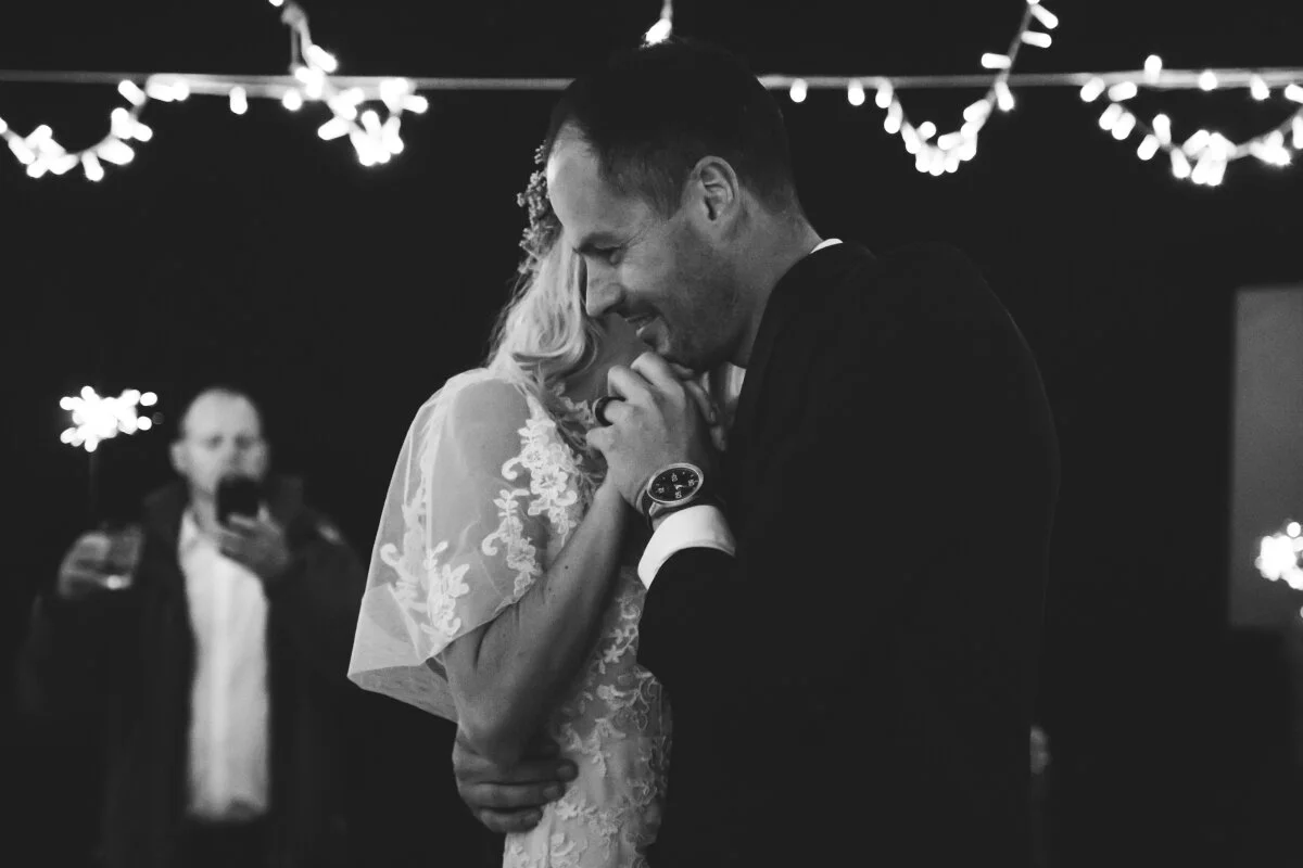 A couple dancing closely at a wedding reception, with string lights hanging in the background, captured in black and white.