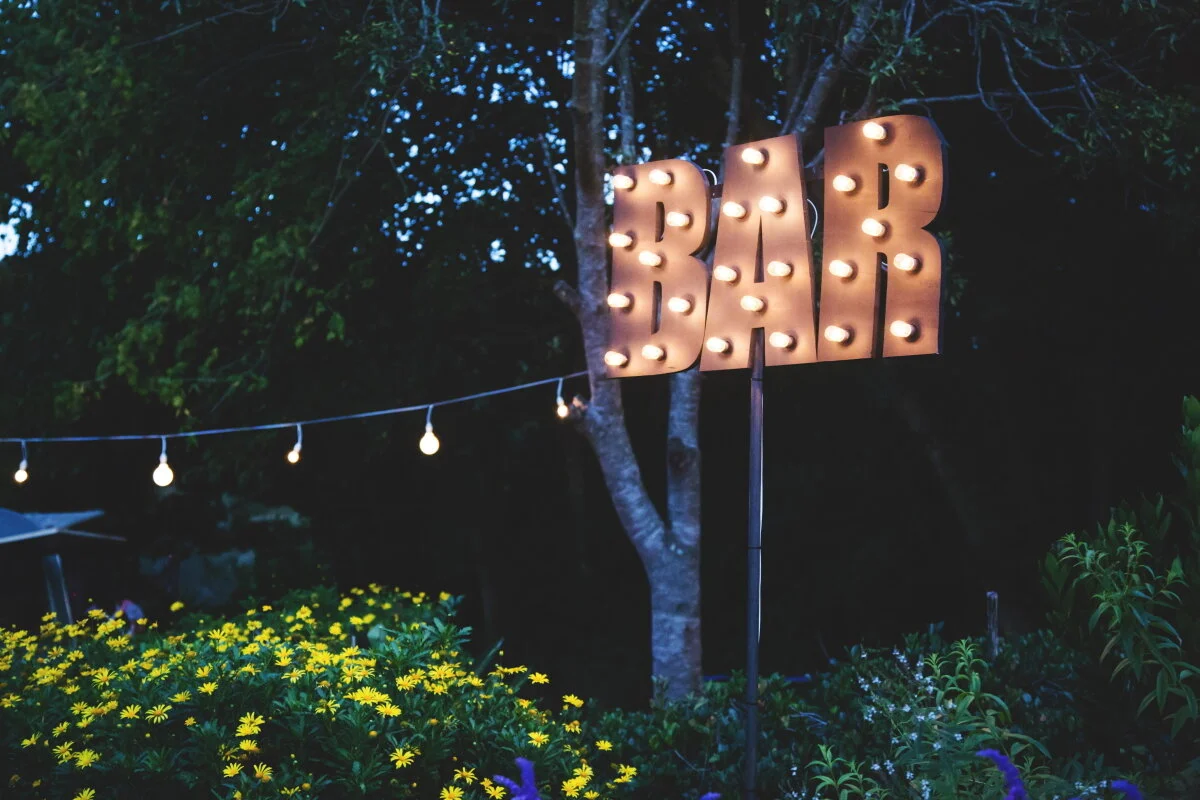 Illuminated sign spelling "BAR" located outdoors at night, surrounded by trees and yellow flowers, with string lights hanging nearby.