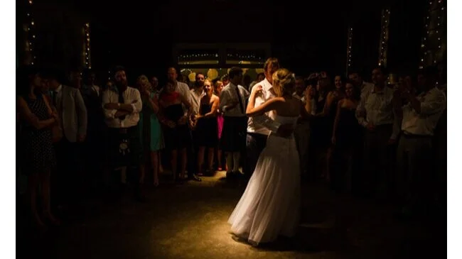A bride and groom share a dance in the center of a dimly lit room, surrounded by guests at a wedding reception.