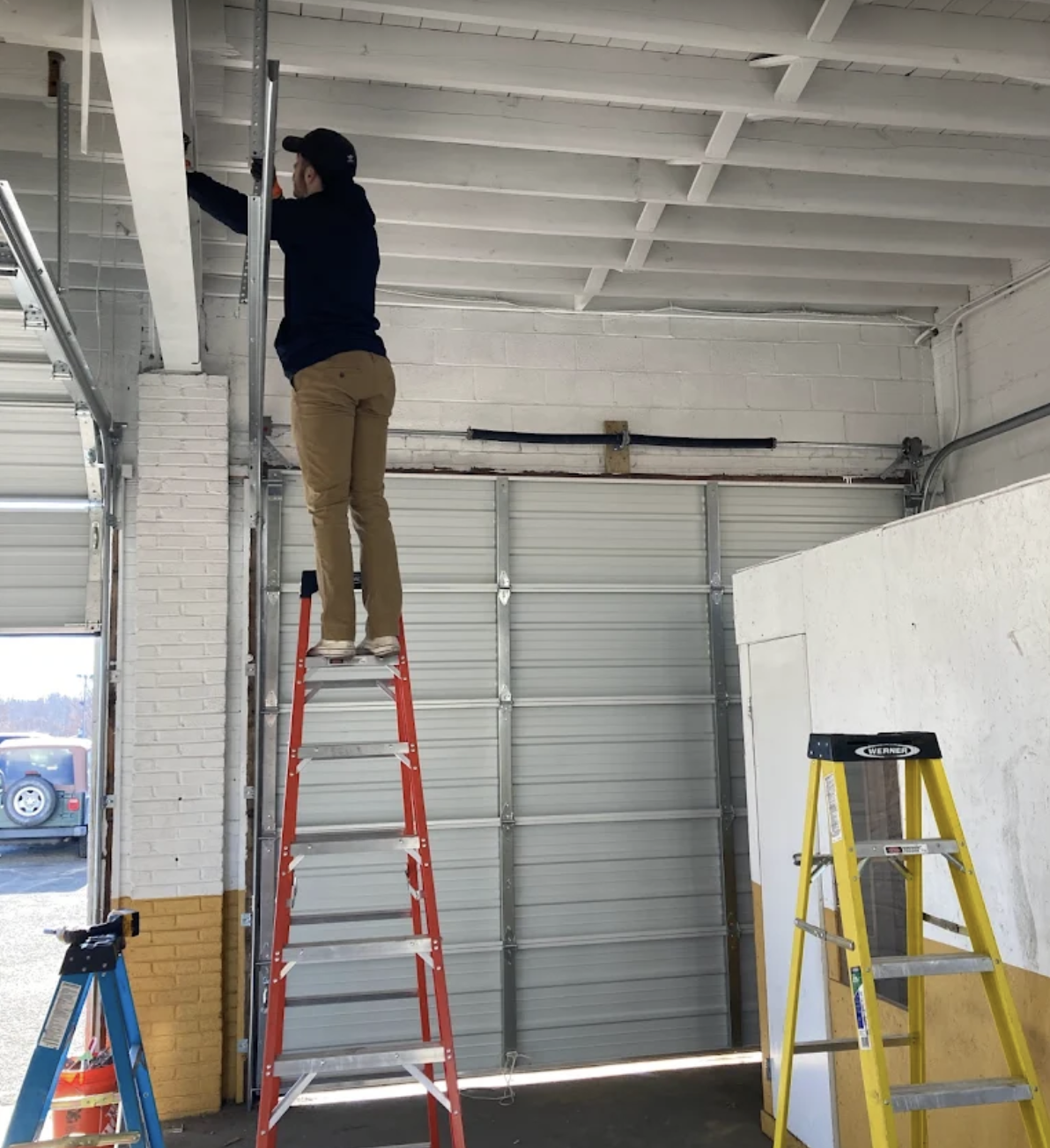 A person working on wiring while standing on a red ladder inside a garage or warehouse, with other ladders and a garage door visible.