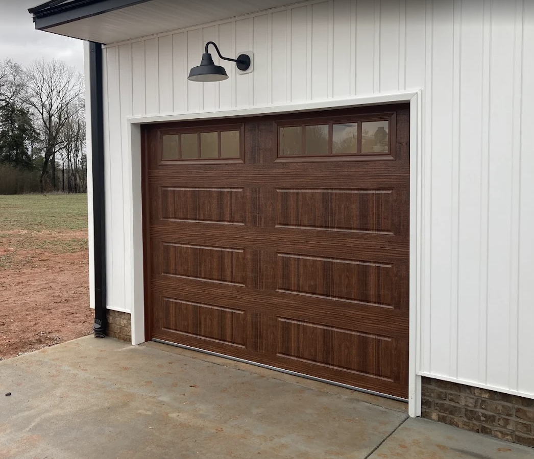 A closed garage door with a dark wood finish and small windows at the top, attached to a white building with vertical siding, outside on a concrete driveway.