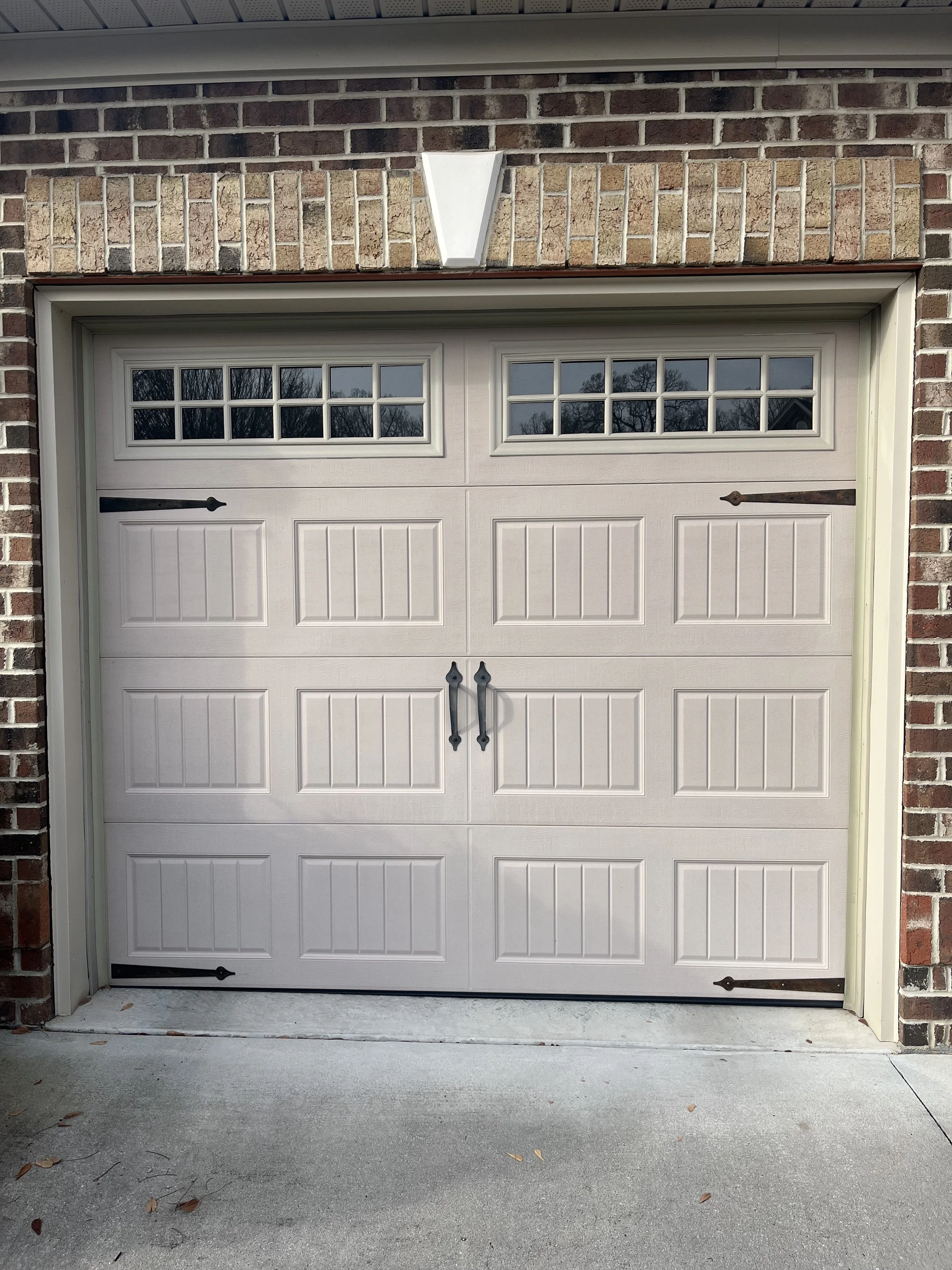 White garage door with black handles and decorative hardware, windows at the top, brick and stone exterior, and a concrete driveway.