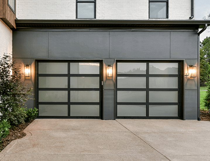 Two modern garage doors with frosted glass panels, flanked by wall-mounted lights, in front of a gray building with a concrete driveway.