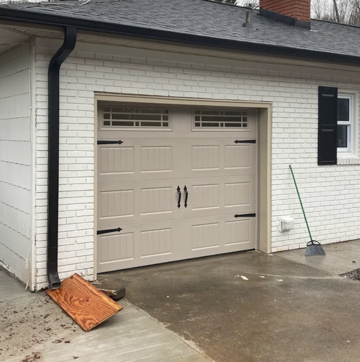 A closed parking garage door on a white brick house with black shutters. A broom leaning against the wall, a wooden board on the ground, and a downspout extending from the roof to the ground.