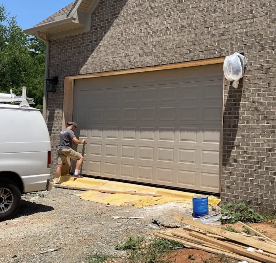 A person working on installing a new garage door. There is construction material and a white van nearby.