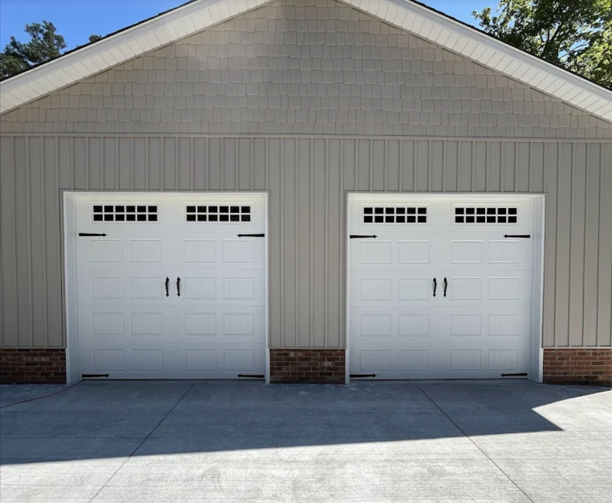 A gray garage with two closed white doors, each having black handles and black decorative hardware, on a concrete driveway with a shadow cast on it.