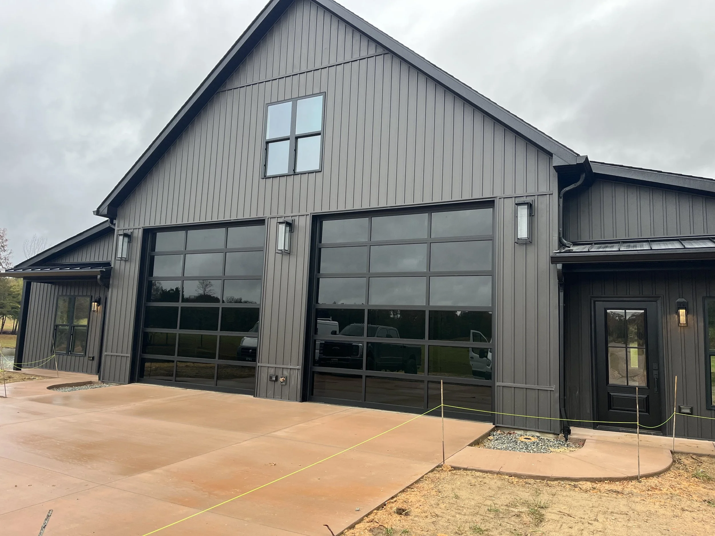 Black modern barn-style building with large glass garage doors, smaller side windows, and a front door, situated on a concrete driveway with a cloudy sky overhead.