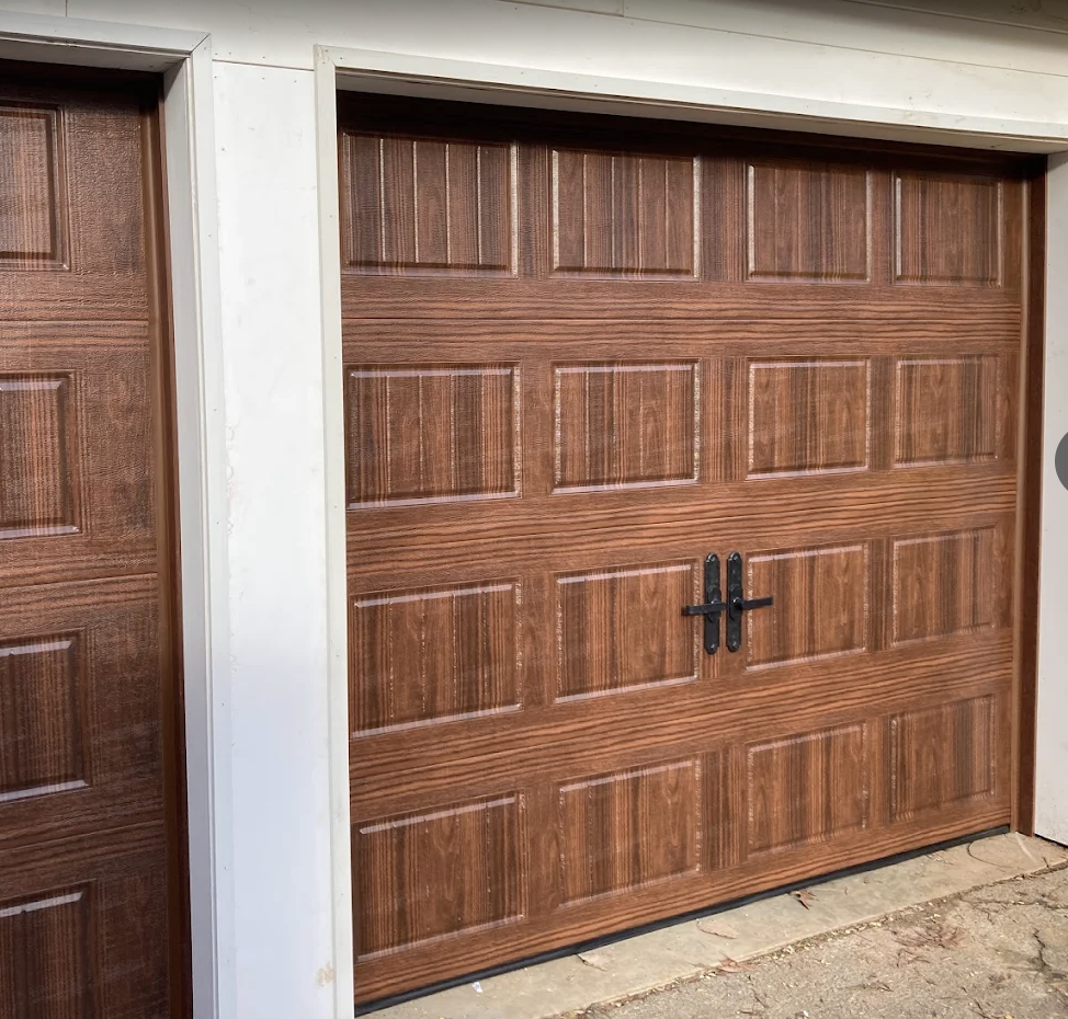 View of a closed brown wooden garage door with black handles, set within a white frame, next to a white wall, on a concrete driveway.