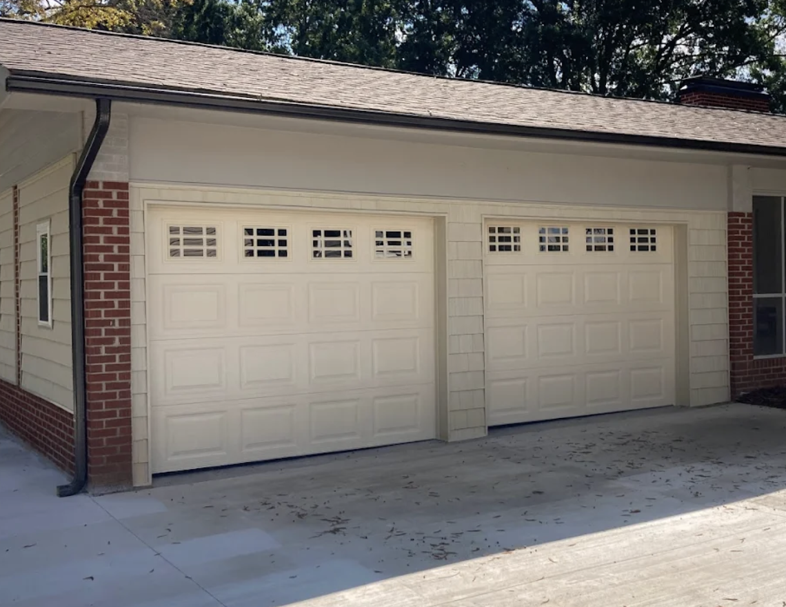 A two-car garage with white doors and small vent windows, attached to a house with beige siding, brick accents, and a shingled roof.
