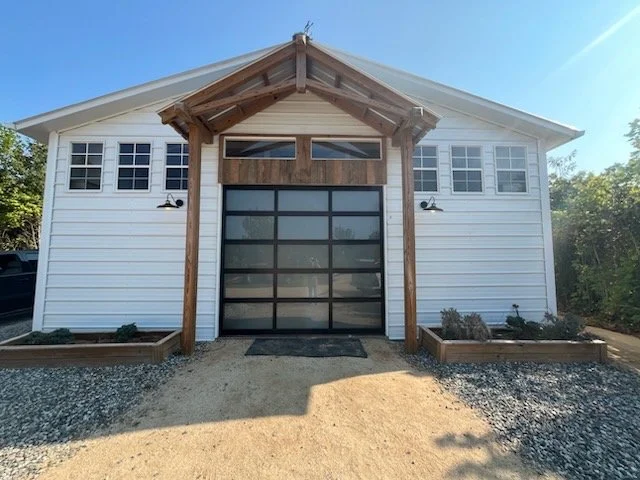 Front view of a white garage with a modern glass door, wood accents, and a small gravel driveway.