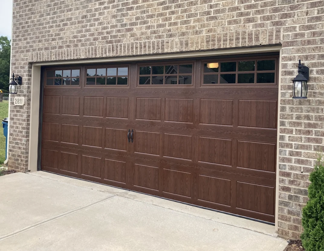 A brown garage door with rectangular panels and small windows at the top, set in a brick house, with black outdoor wall lanterns on either side.