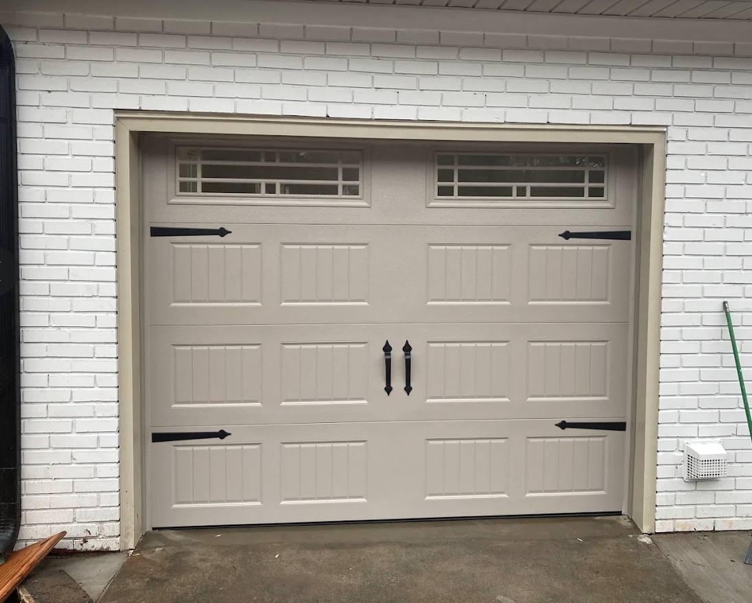 A beige garage door with black hardware, located in a white brick wall with a window near the top. A broom leans against the wall on the right side, and a small object is attached to the wall near the ground on the right.