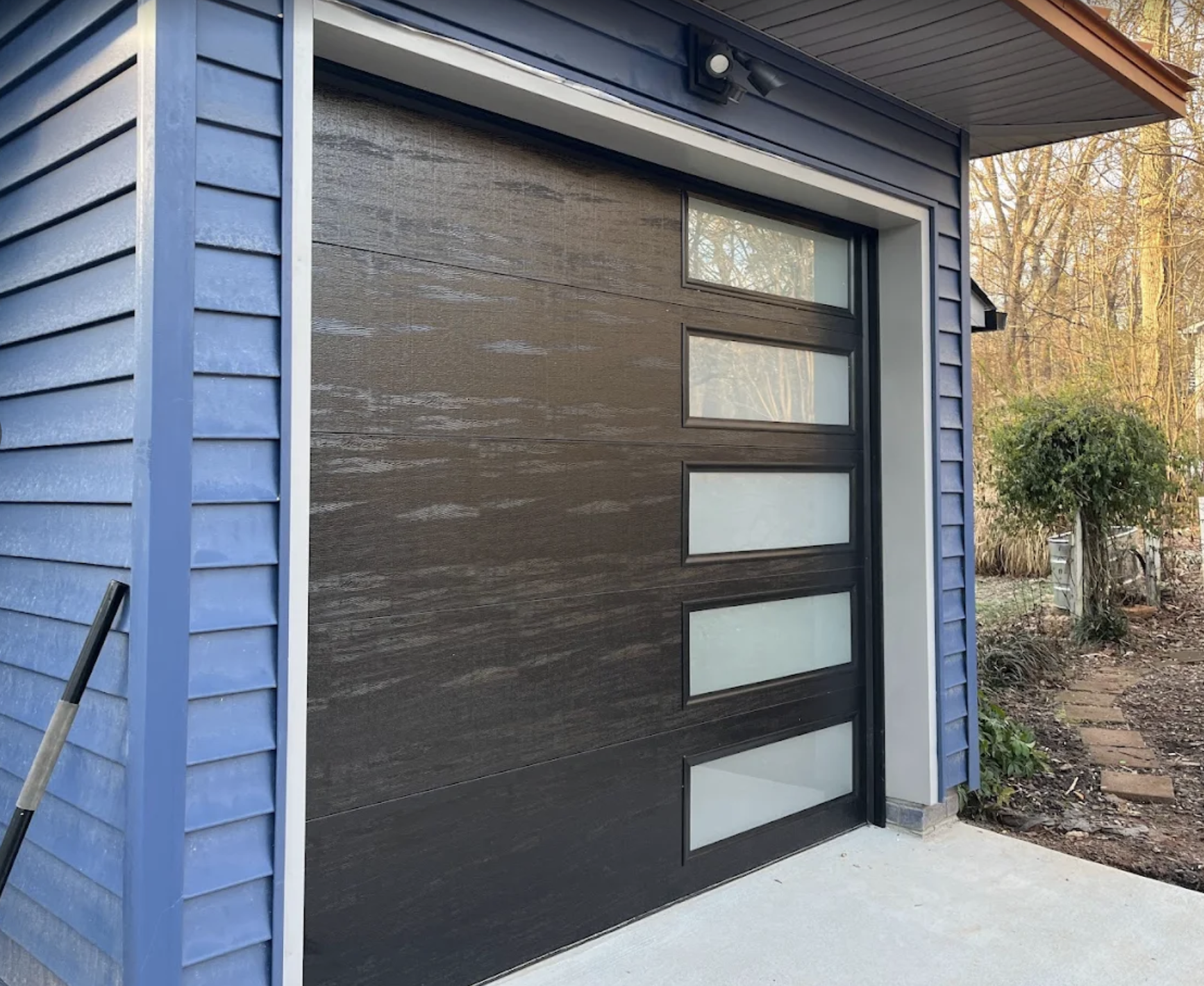 Modern garage door with four horizontal frosted glass panels, set in a house with blue-painted exterior siding, surrounded by a garden with trees and plants.